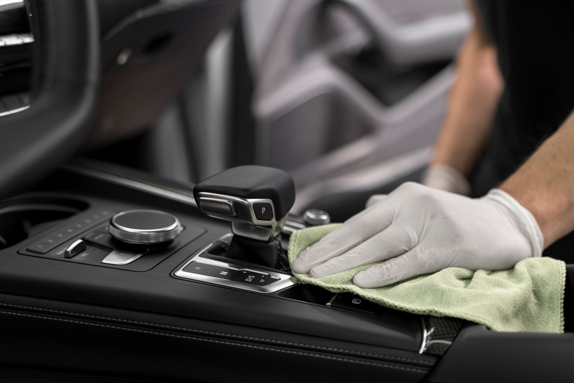 A gloved hand uses a green cloth to clean the center console and gear shifter of a vehicle interior.