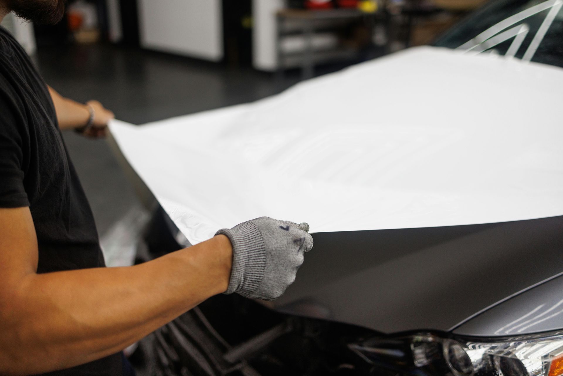 A person wearing a grey glove applies a white adhesive film to the hood of a grey car in a garage.