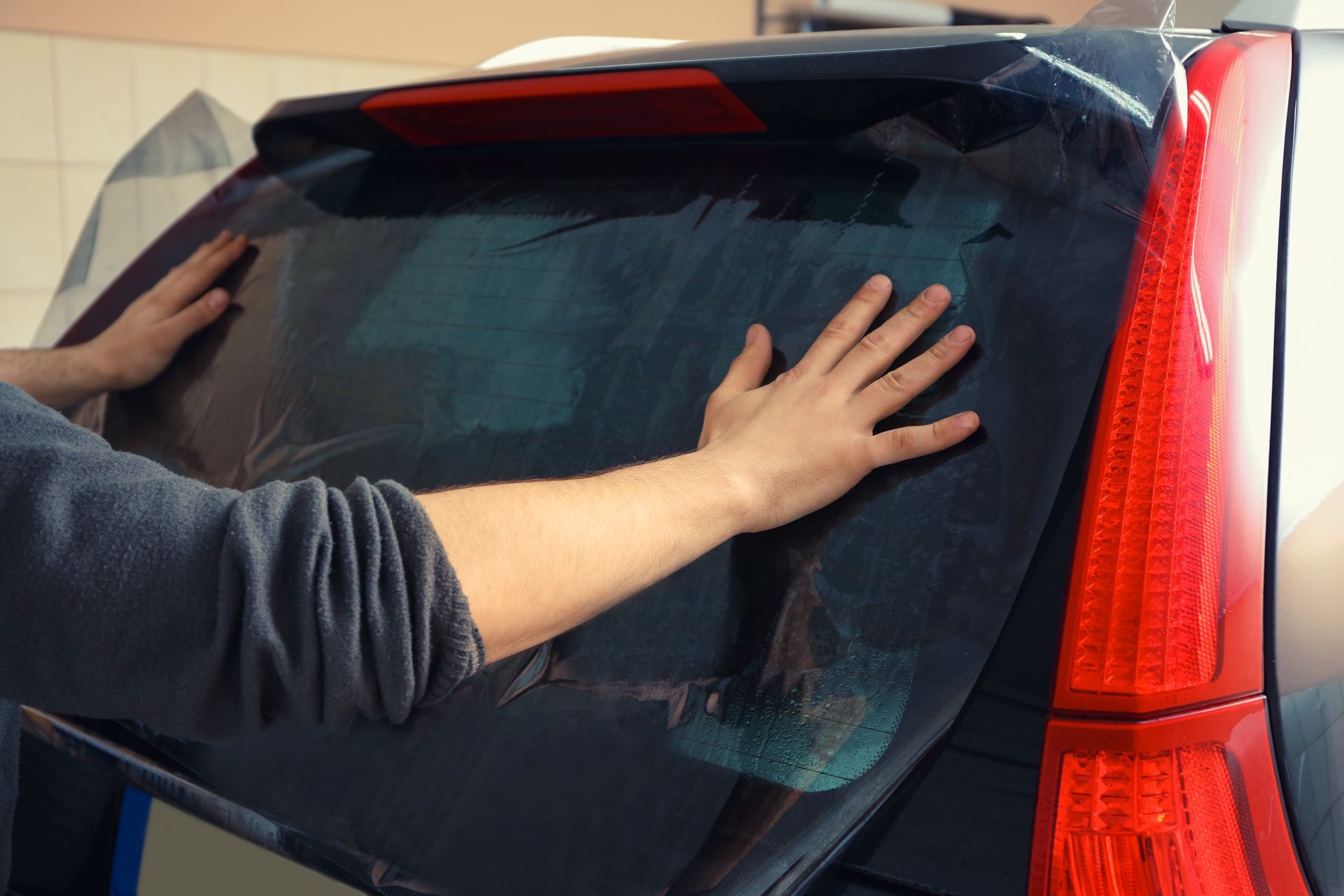 A technician applies a dark tint film to the rear window of a car, smoothing it out with their hands.