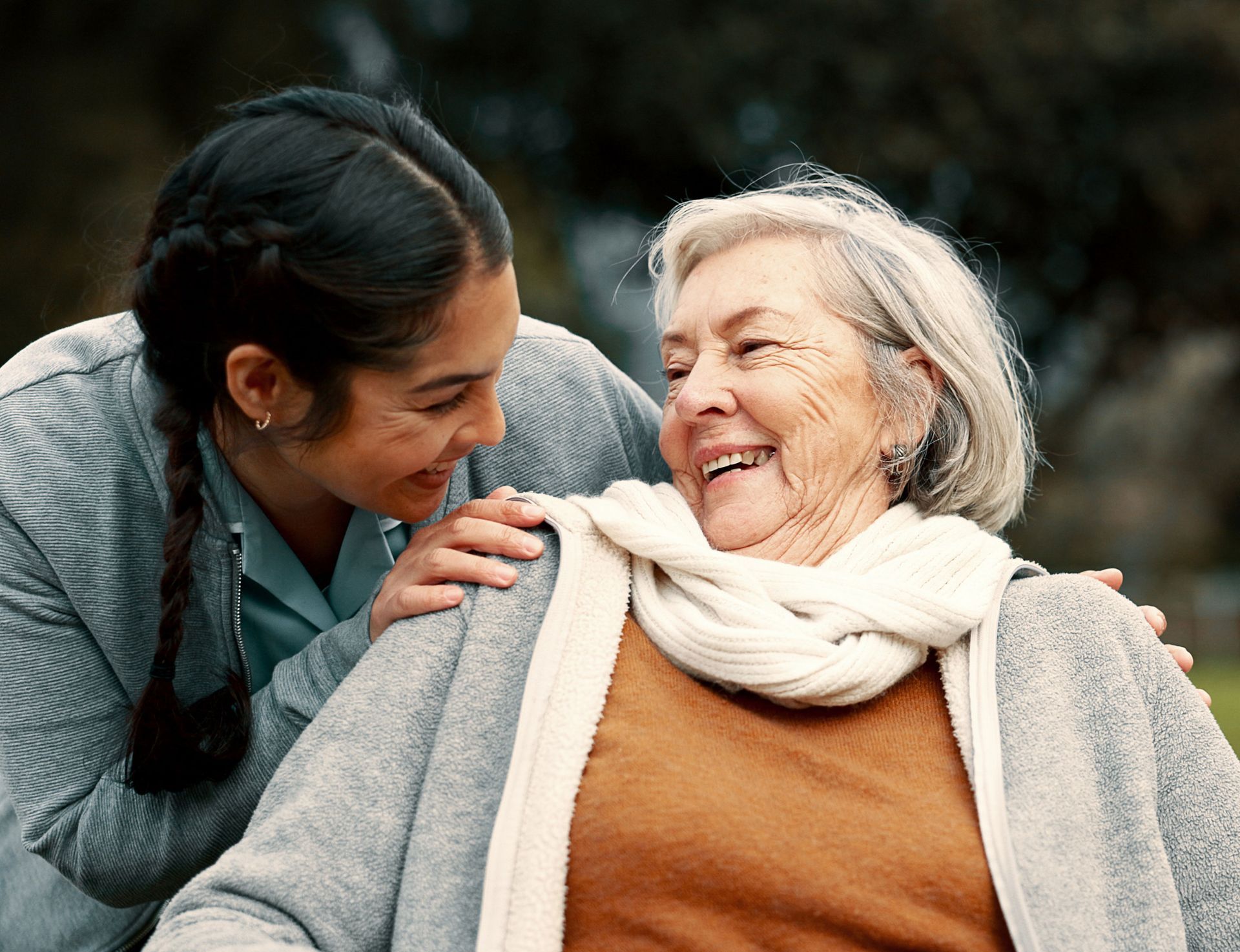 Young woman smiles, placing hands on the shoulders of an older woman, both looking at each other outdoors.