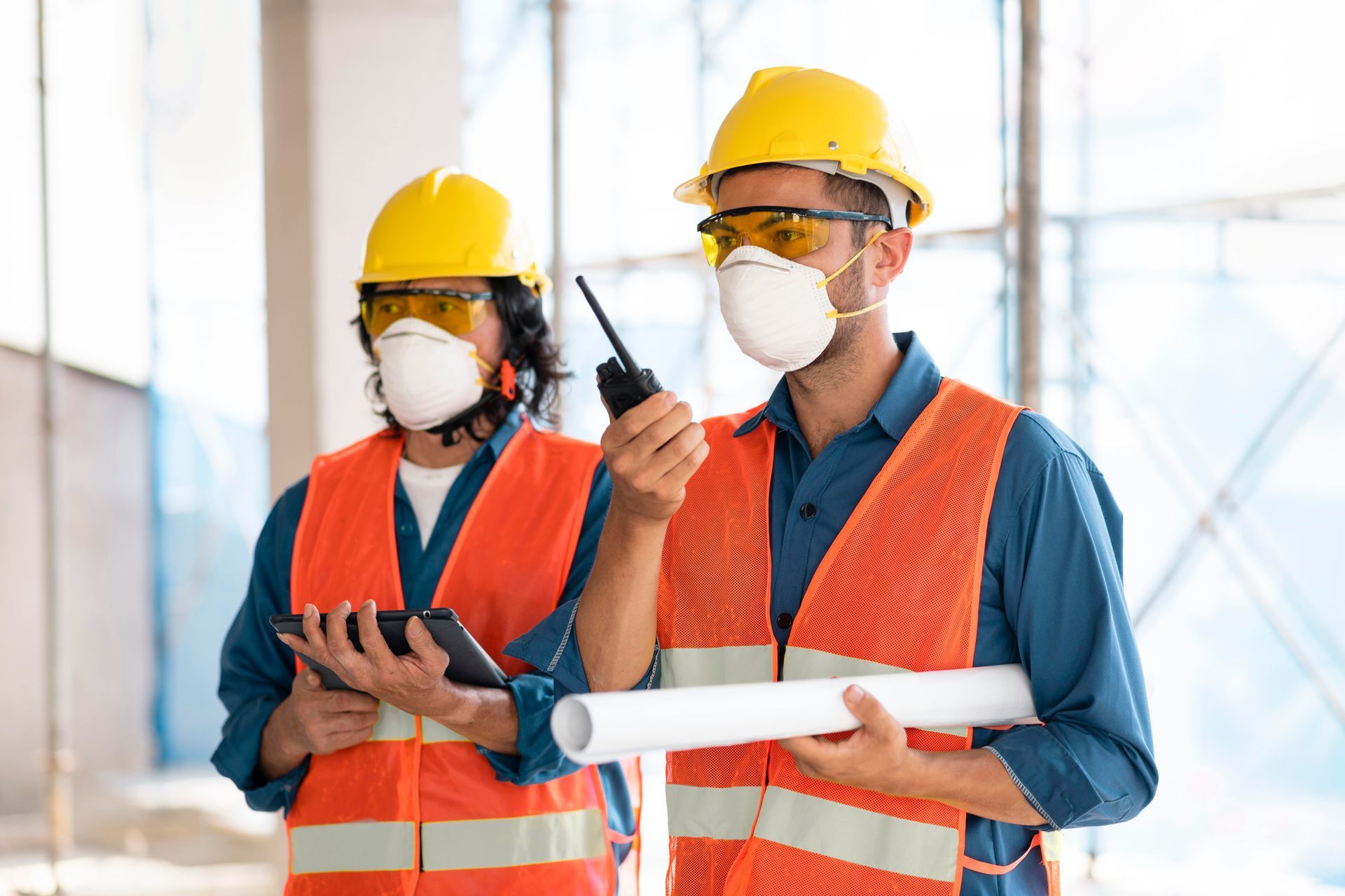 Two construction workers wearing masks and safety vests are standing next to each other.