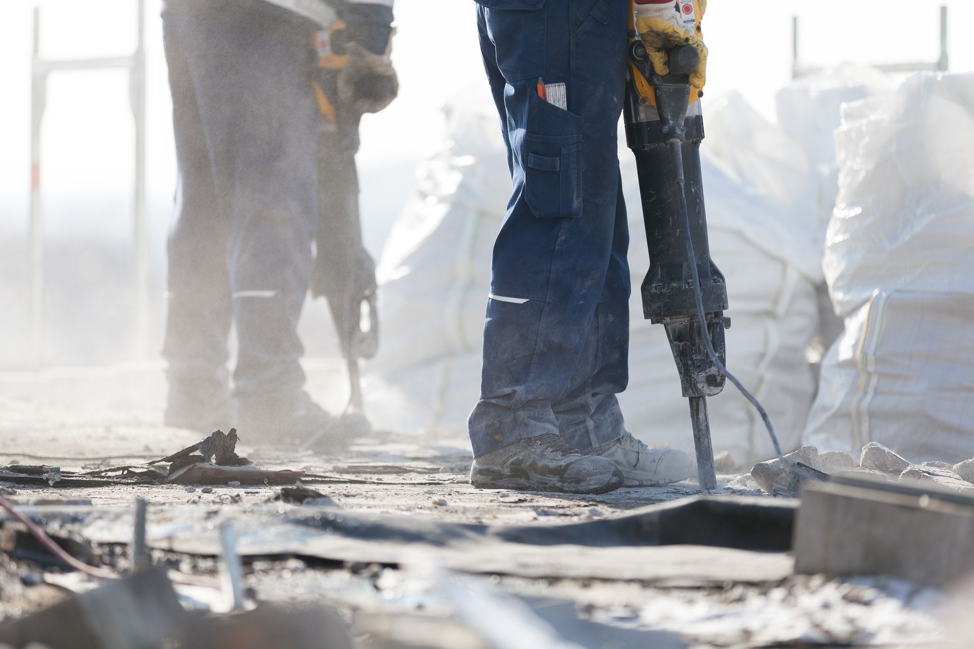 Des ouvriers du bâtiment, vêtus de pantalons et de bottes de travail, manient des marteaux-piqueurs sur un chantier poussiéreux et jonché de débris.