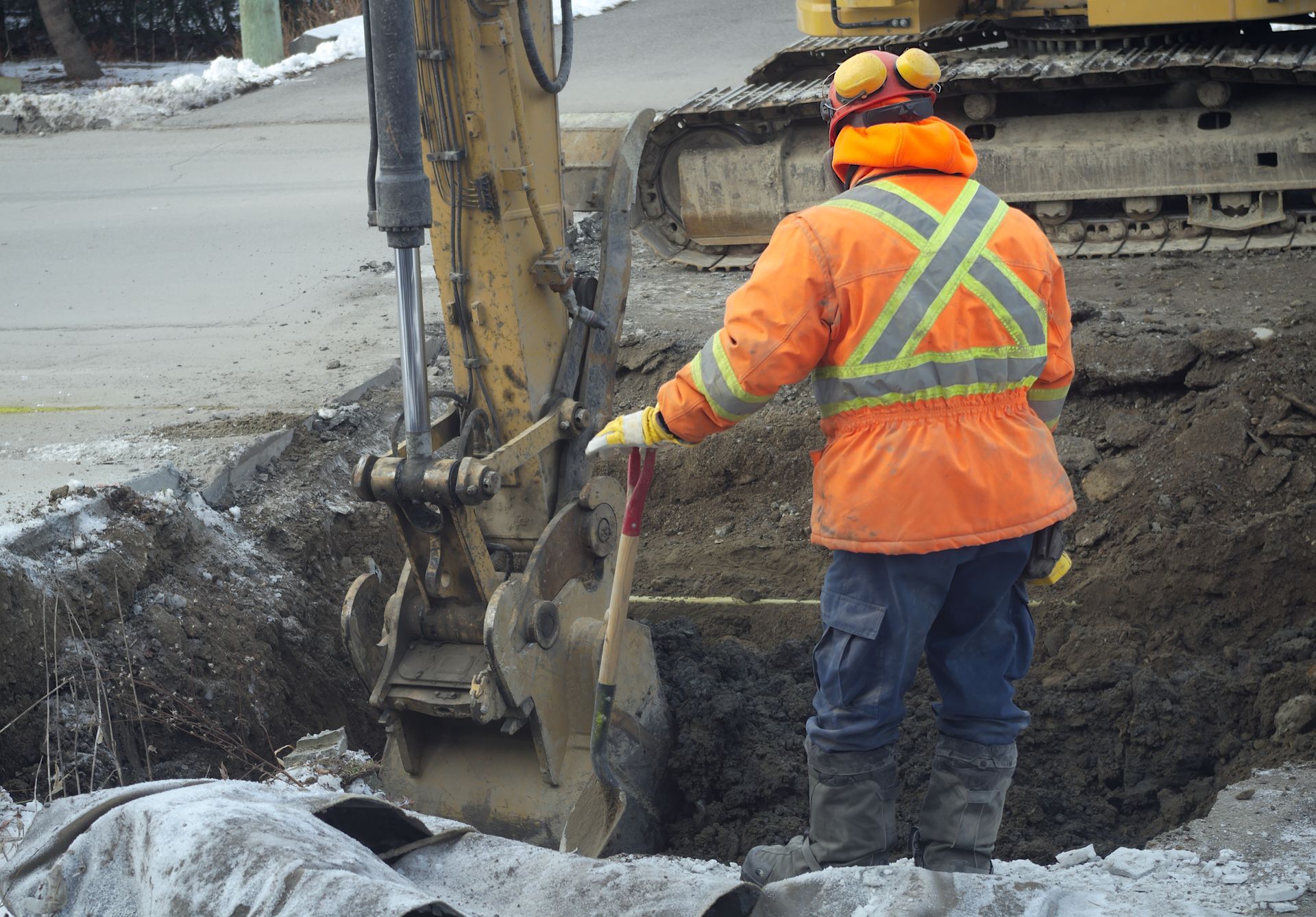 Un ouvrier du bâtiment vêtu d'une veste orange haute visibilité se tient dans une fosse d'excavation près d'une machinerie jaune.
