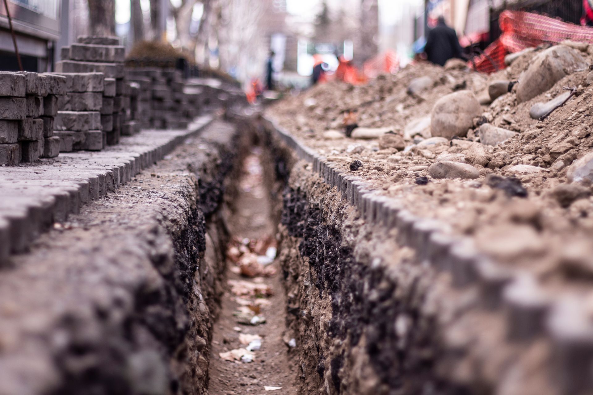 Une tranchée de chantier étroite et profonde traverse une rue urbaine, bordée de piles de pavés et de tas de terre.
