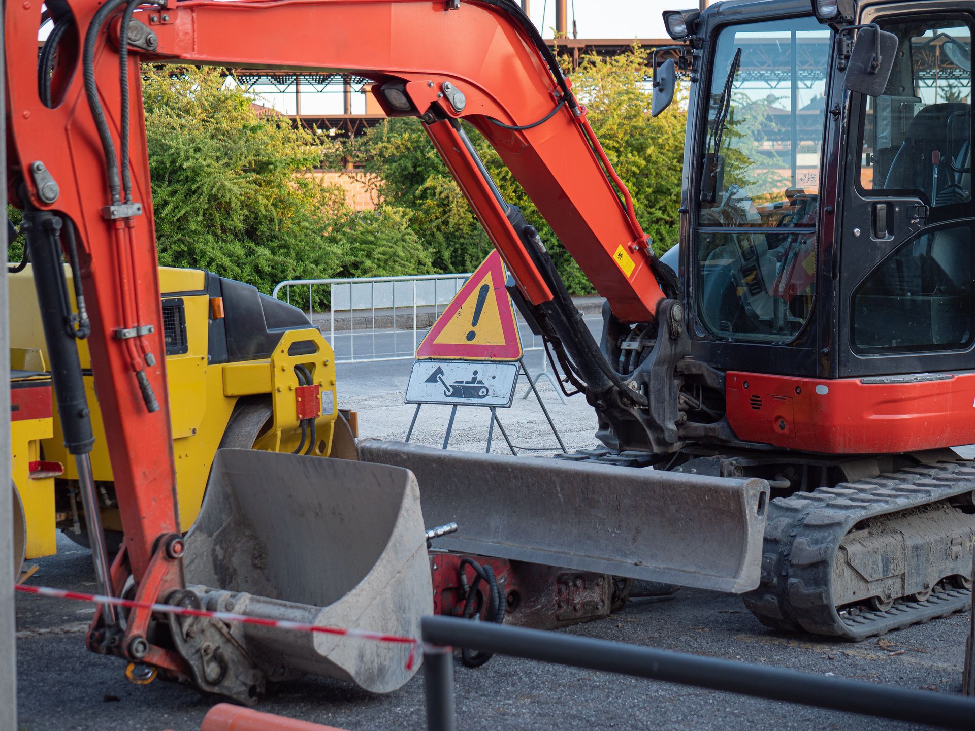Une pelleteuse orange garée à côté d'un rouleau compresseur jaune, avec un panneau de signalisation triangulaire sur un chantier.