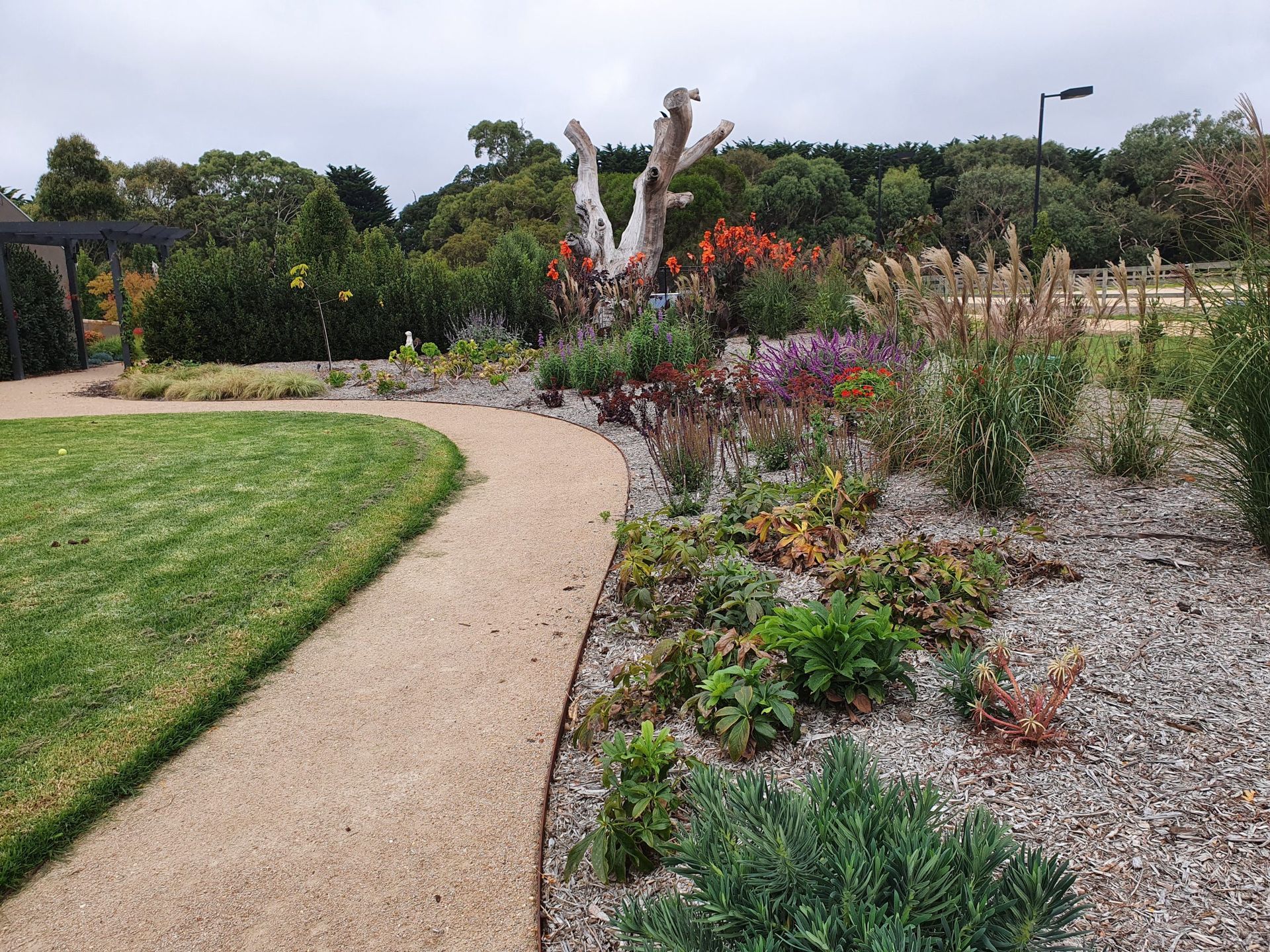 A winding gravel path borders a vibrant flower bed with a weathered tree sculpture, set against a green lawn and trees.