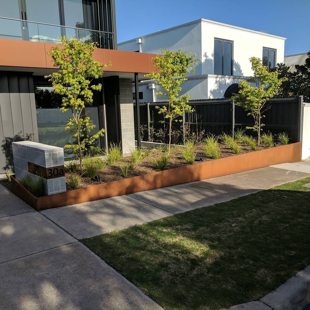 Rusty metal planter box with young trees and greenery in front of a modern building and white house.