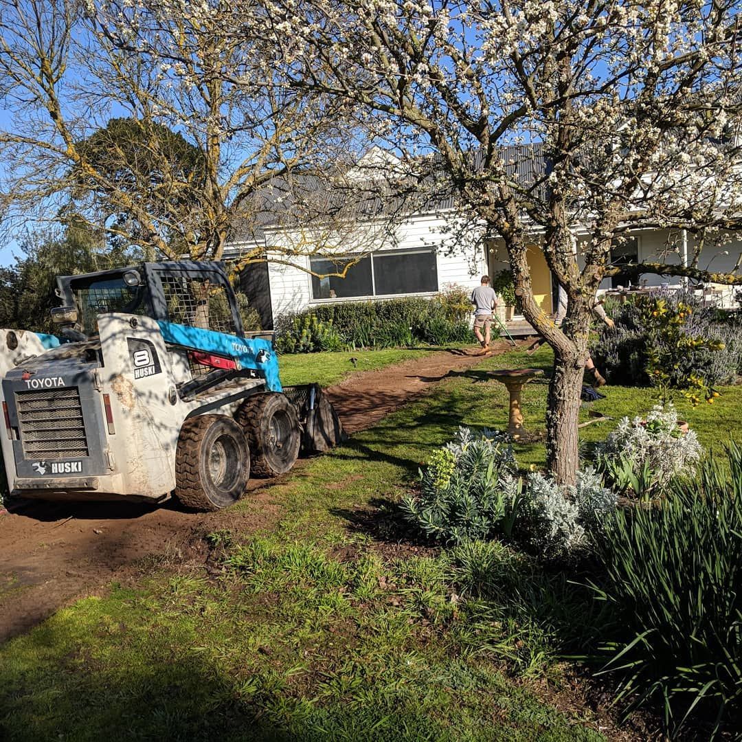 A skid-steer loader parked on a muddy lawn next to a house with a person standing nearby.