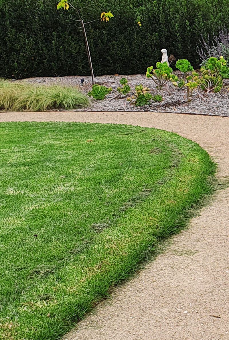 Curved green lawn borders a tan gravel path; background includes greenery and trees.