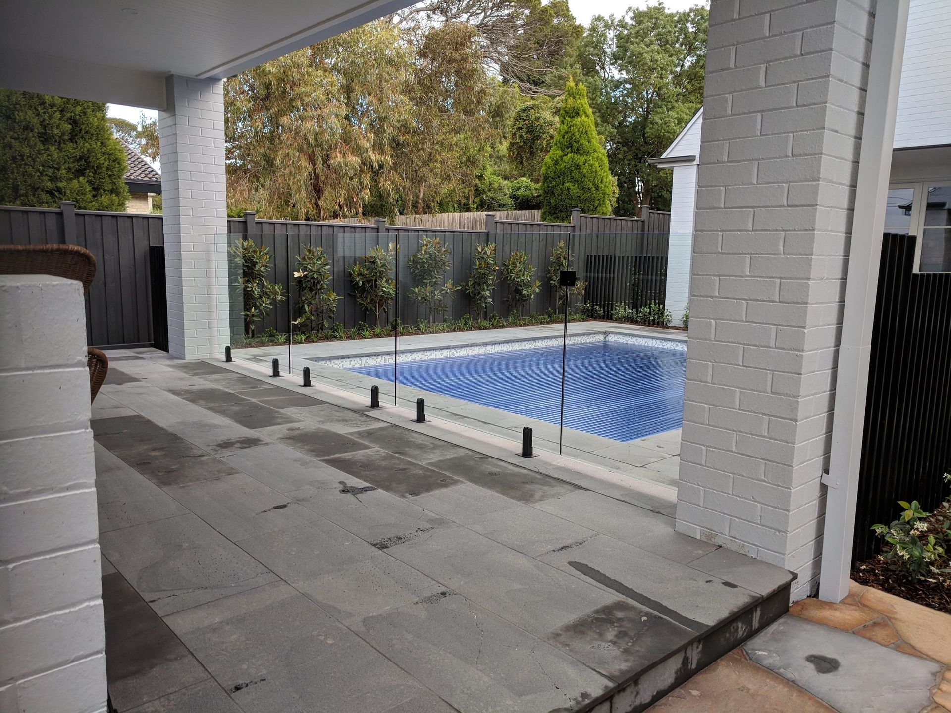 Patio with gray stone tiles, pool with blue water, and glass fence surrounded by trees.