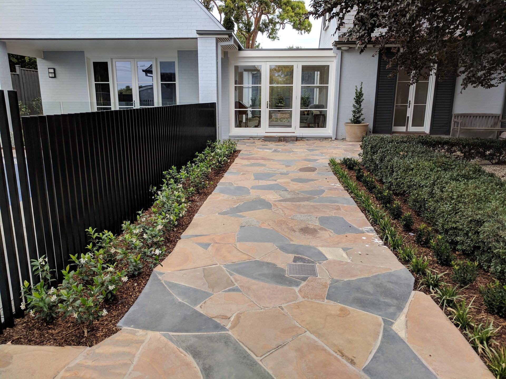 Stone path leading to a house with a white door and windows, flanked by green hedges and a black fence.