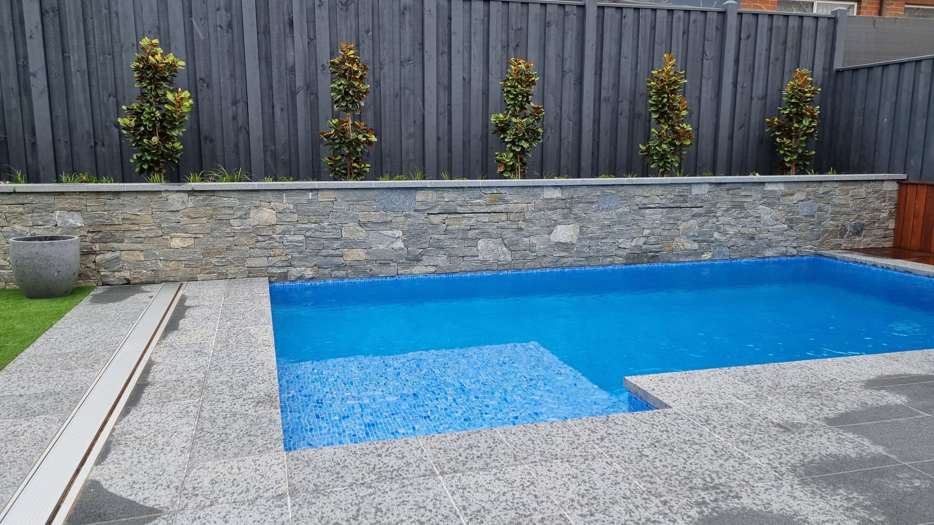 Swimming pool with blue water and stone wall, bordered by landscaping and a wooden fence.