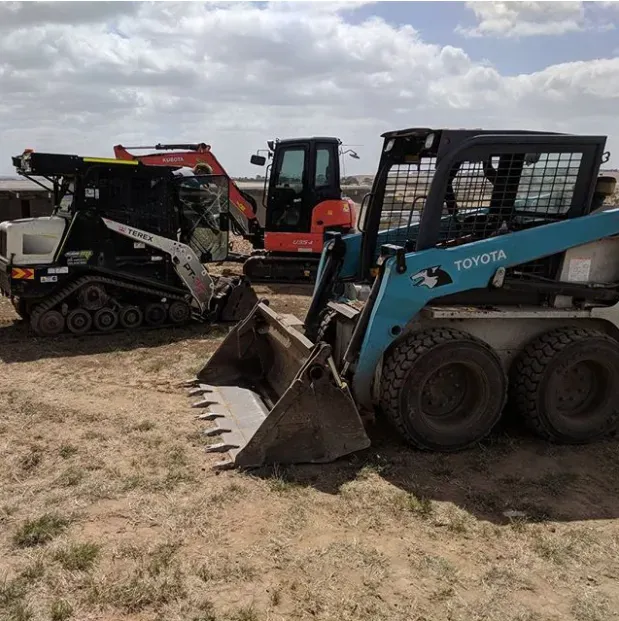 Three construction vehicles, including a blue Toyota skid steer, on a dirt field under a cloudy sky.