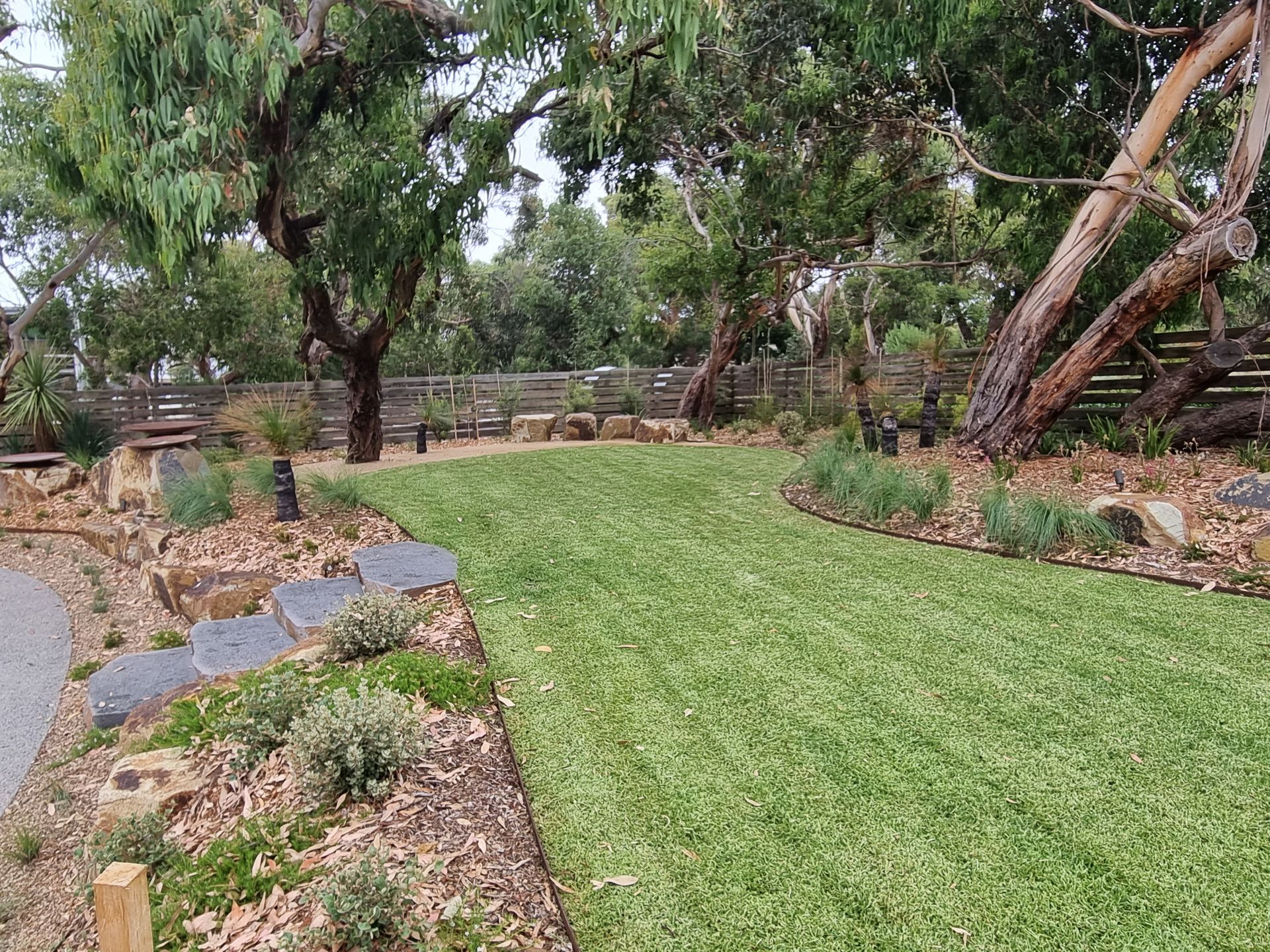 Grassy path through a garden with trees, rock borders, and a wooden fence in the background.