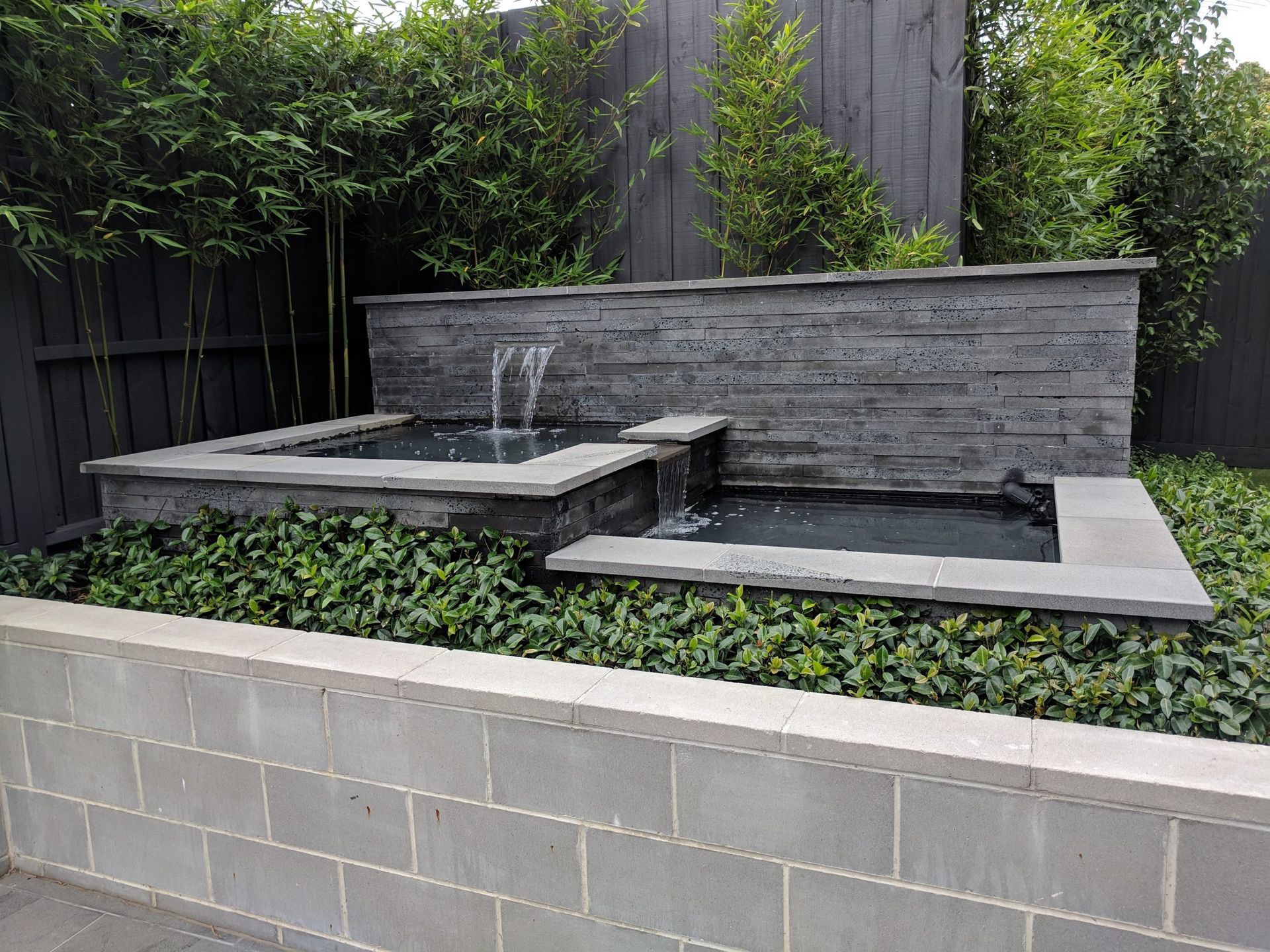 Water feature with tiered pools, gray stonework, and greenery against a dark wooden fence.