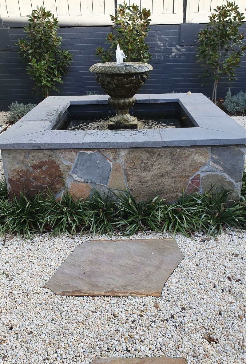 Stone fountain in a square stone basin with water feature in gravel-filled garden.
