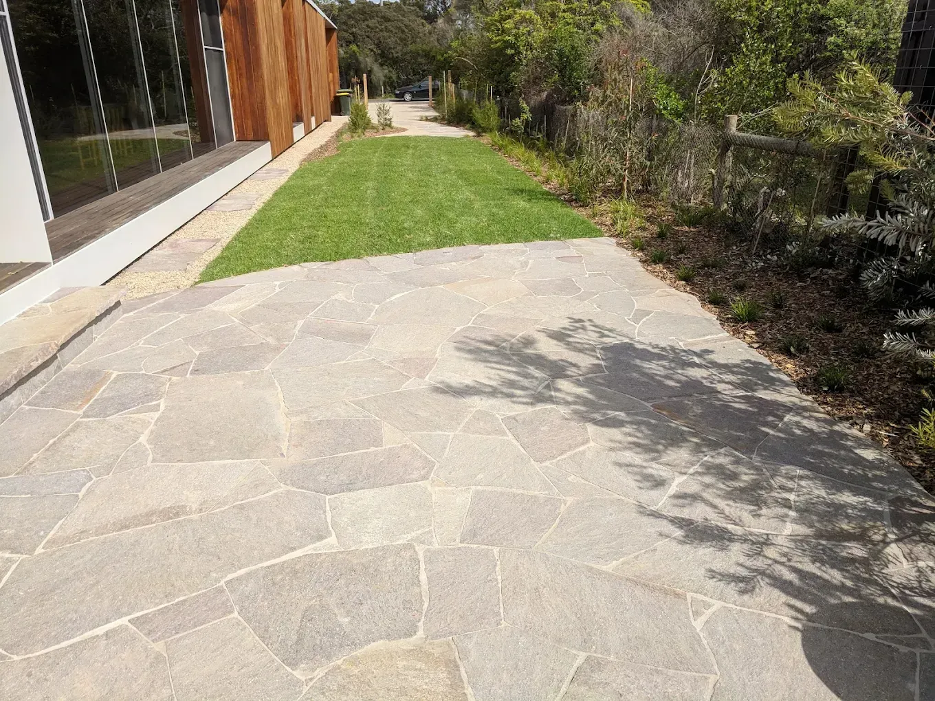 Flagstone path leads towards a grassy area and building with wooden siding, next to a landscaped garden.