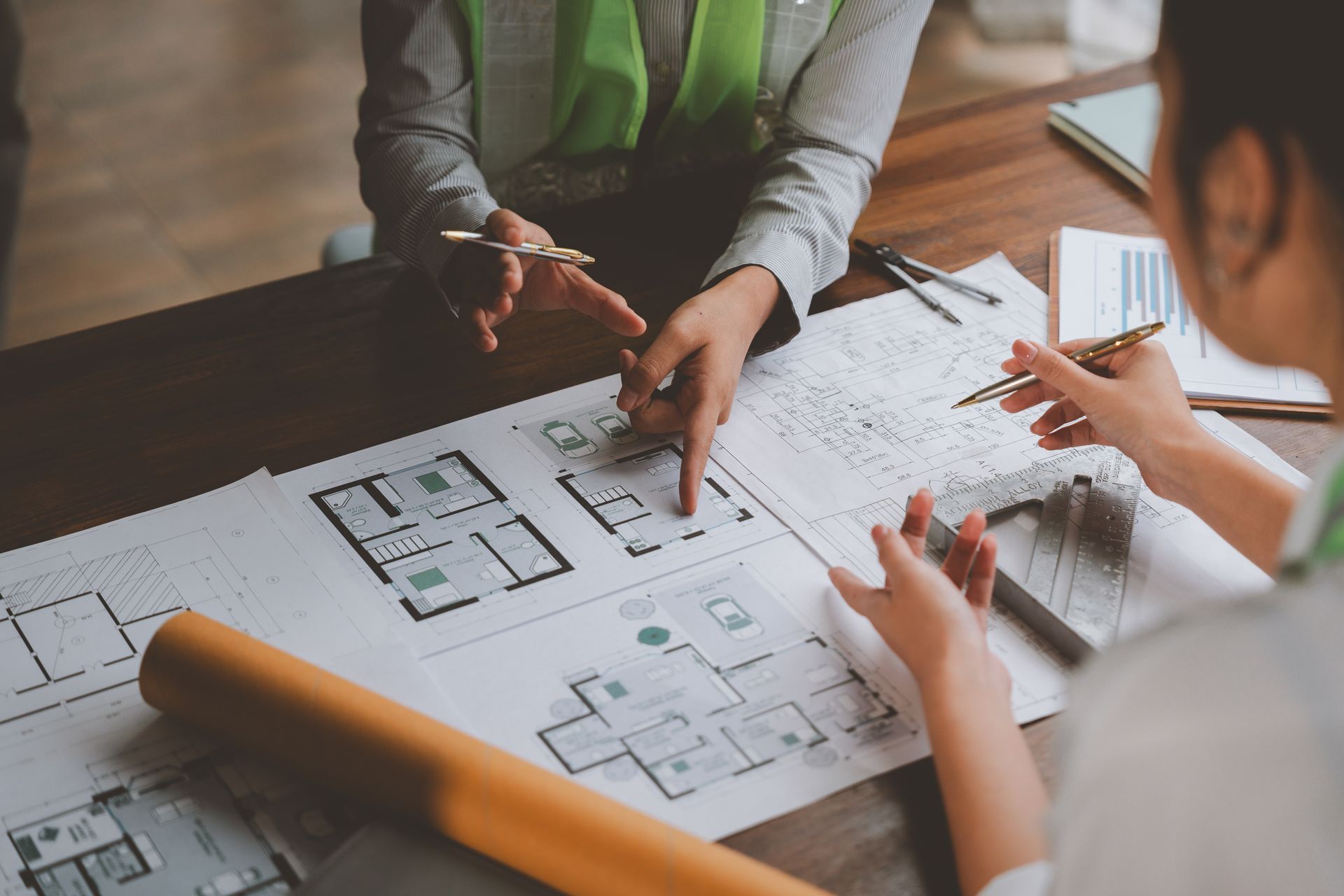 Two people reviewing architectural blueprints at a desk, pointing, with papers and a rolled-up plan.