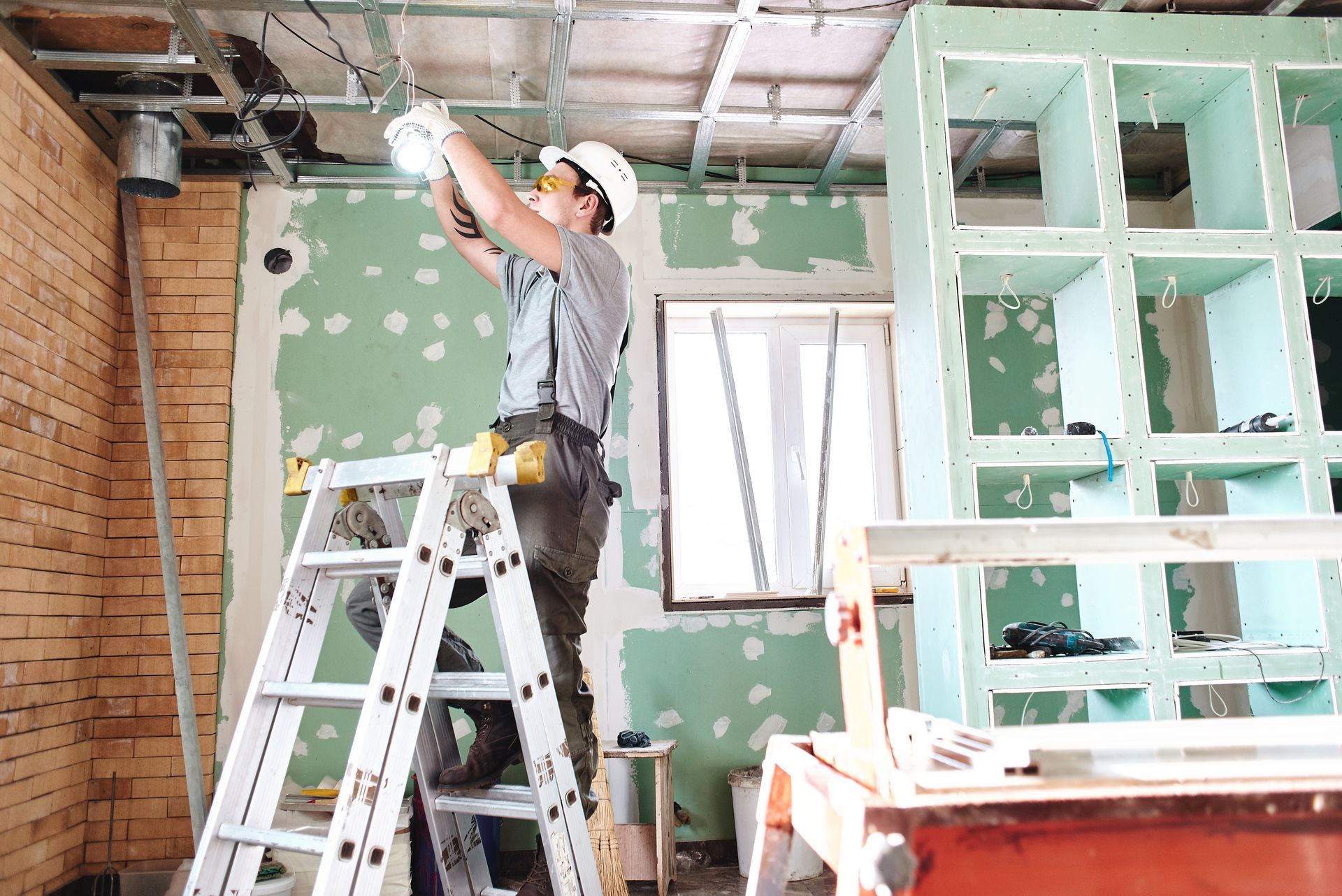 Construction worker on a ladder installing wiring in a room under renovation; unfinished walls and ceiling.