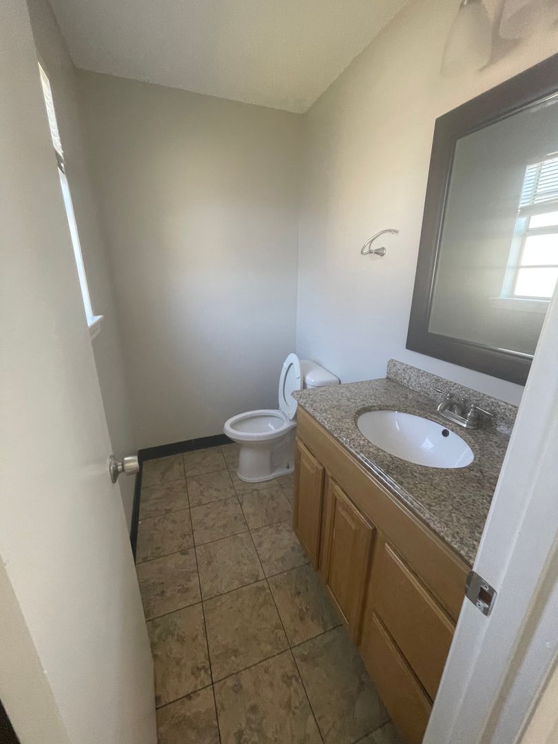 Bathroom with toilet, sink, mirror, and wooden vanity. Tile floor, white door and gray walls.
