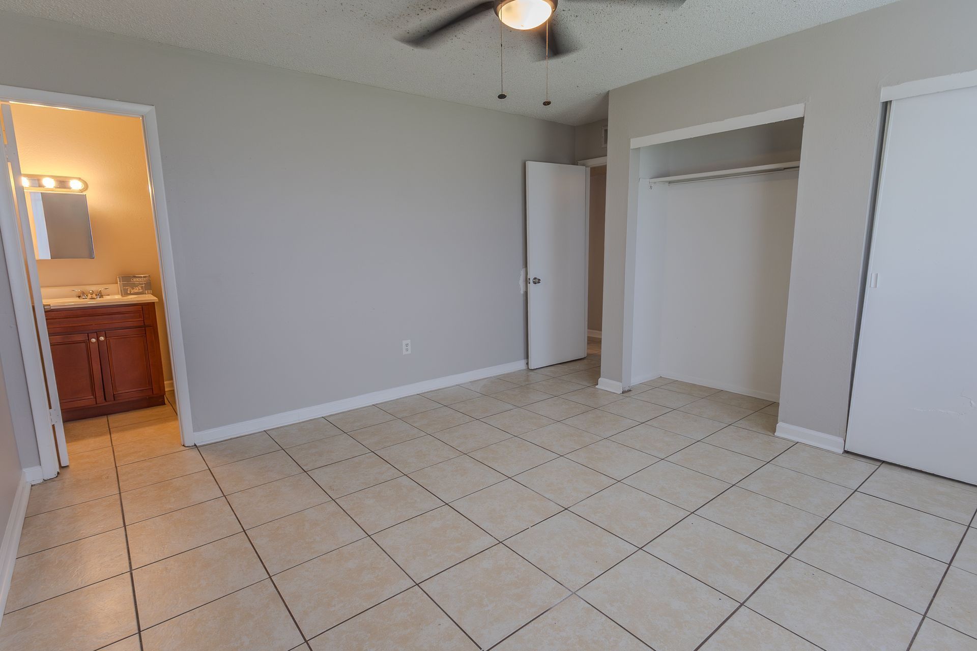 Empty bedroom with tile floor, closet, open door to bathroom with vanity. Gray walls, ceiling fan.