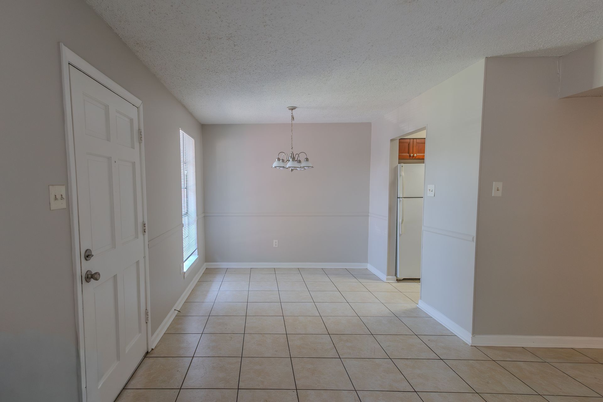 Empty dining room and kitchen with white door, light walls, and tiled floors.