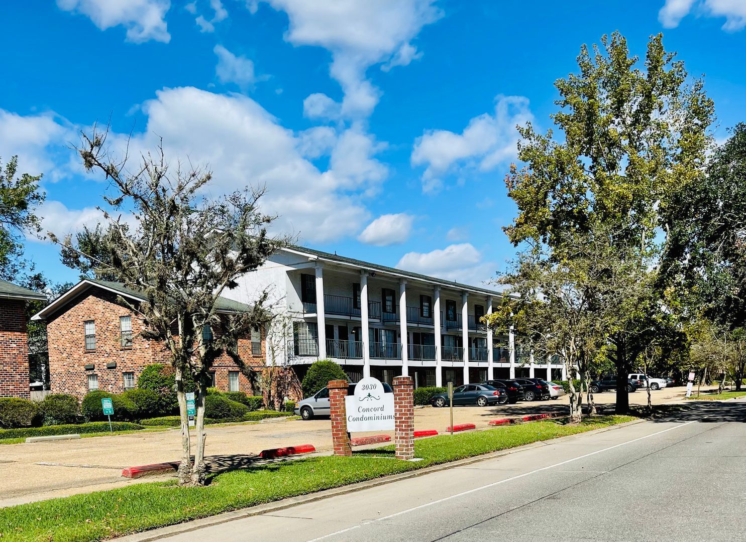Apartment building with balconies and a brick facade, trees, cars, and a sign on a sunny day.