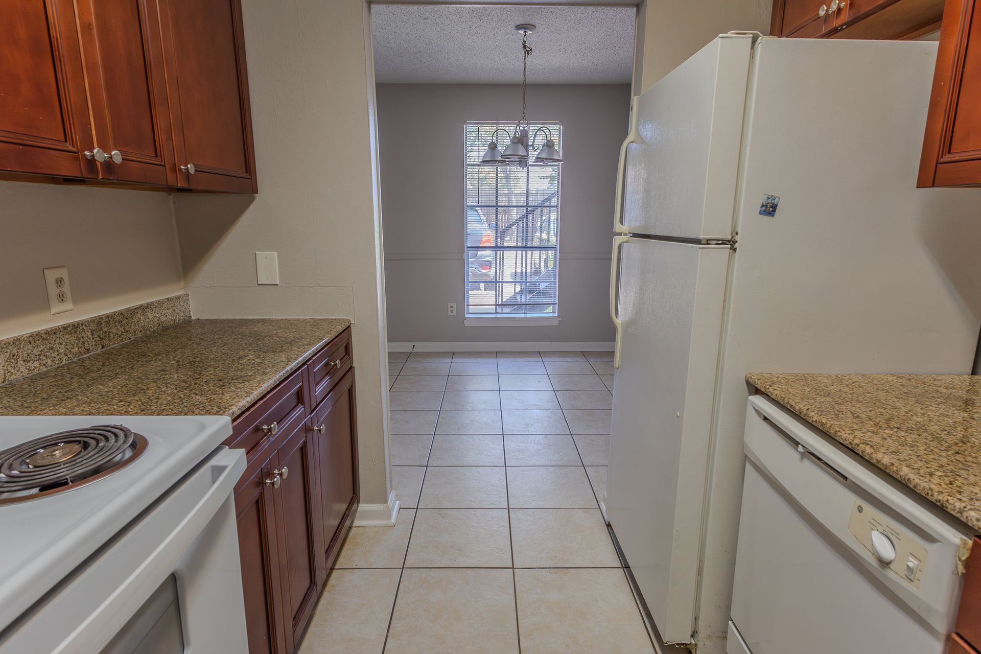 Kitchen with brown cabinets, white appliances, and a window at the end of a hallway.