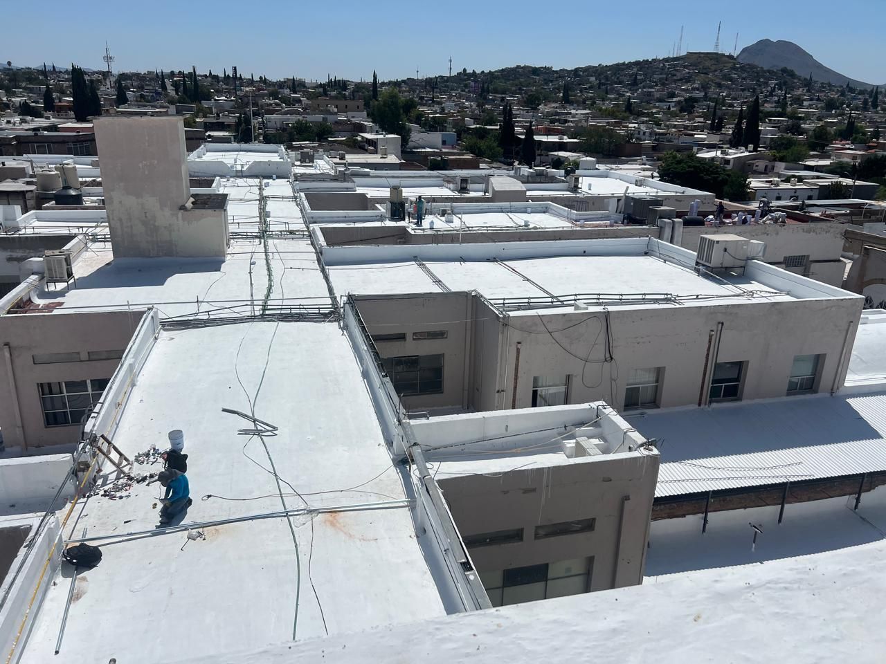 Vista de tejados con superficies blancas; una persona trabaja cerca de un equipo. Ciudad y colinas al fondo.
