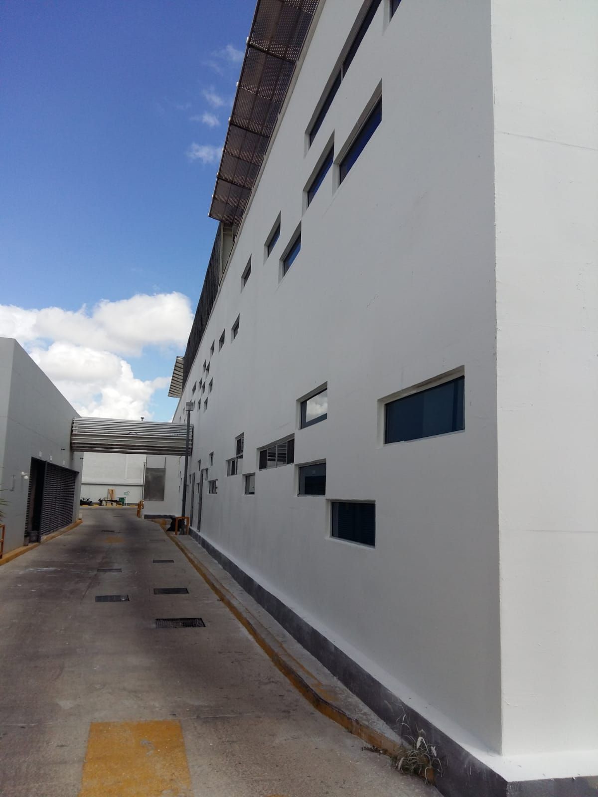 Edificio industrial blanco con pequeñas ventanas rectangulares a lo largo de un callejón estrecho. Cielo azul y nubes.