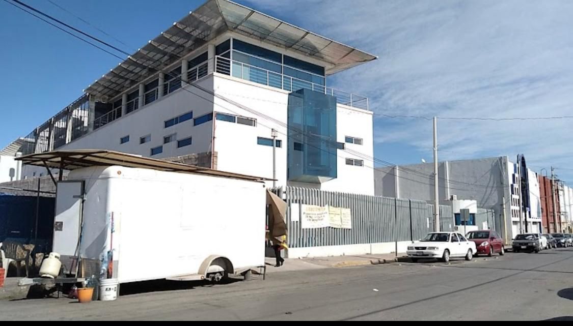 Edificio comercial blanco con detalles azules, carrito de comida al frente, autos en la calle y cerca gris.