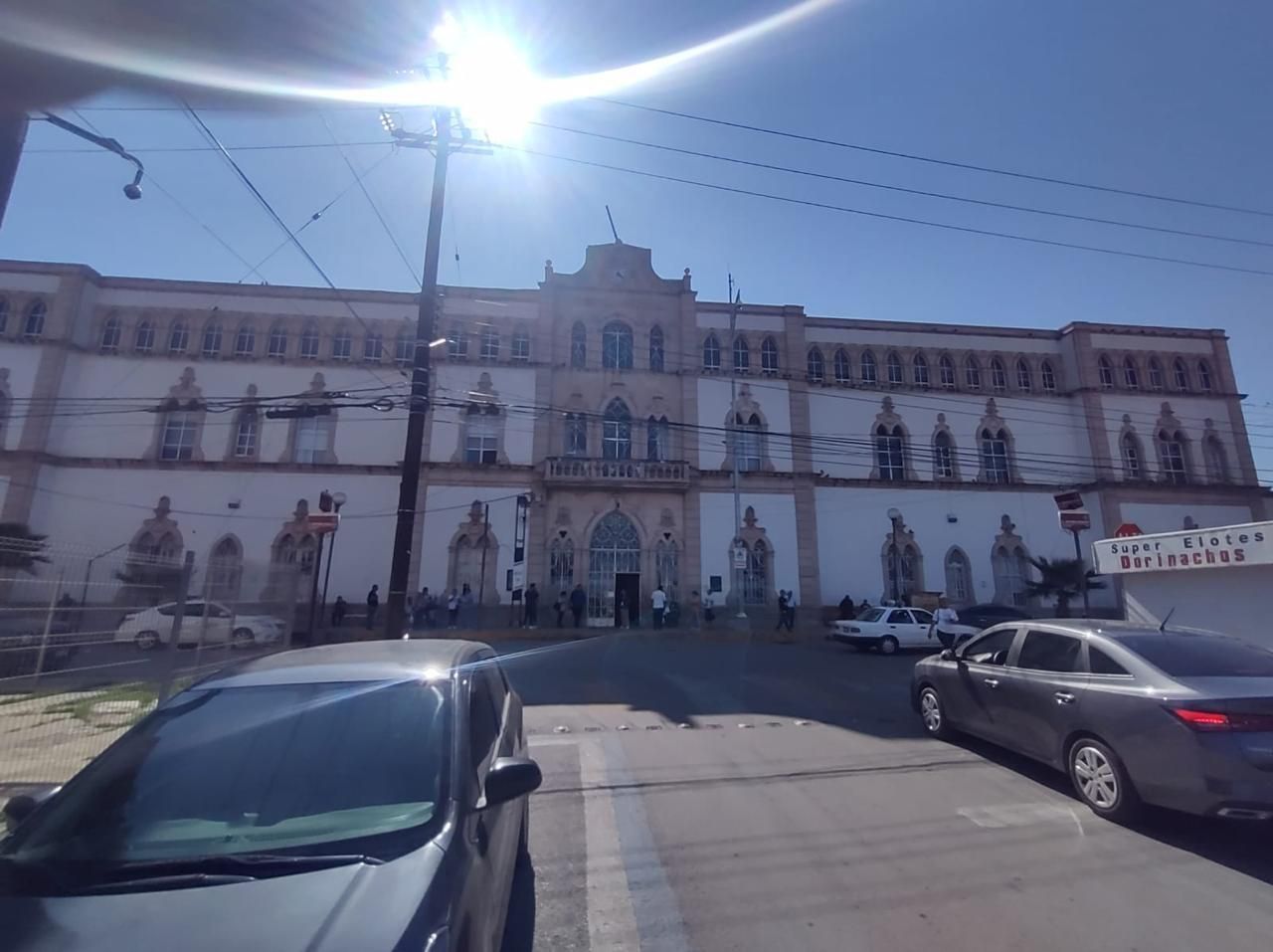 Edificio grande, de color claro, con fachada ornamentada, vista a la calle con autos y cables aéreos, día soleado.
