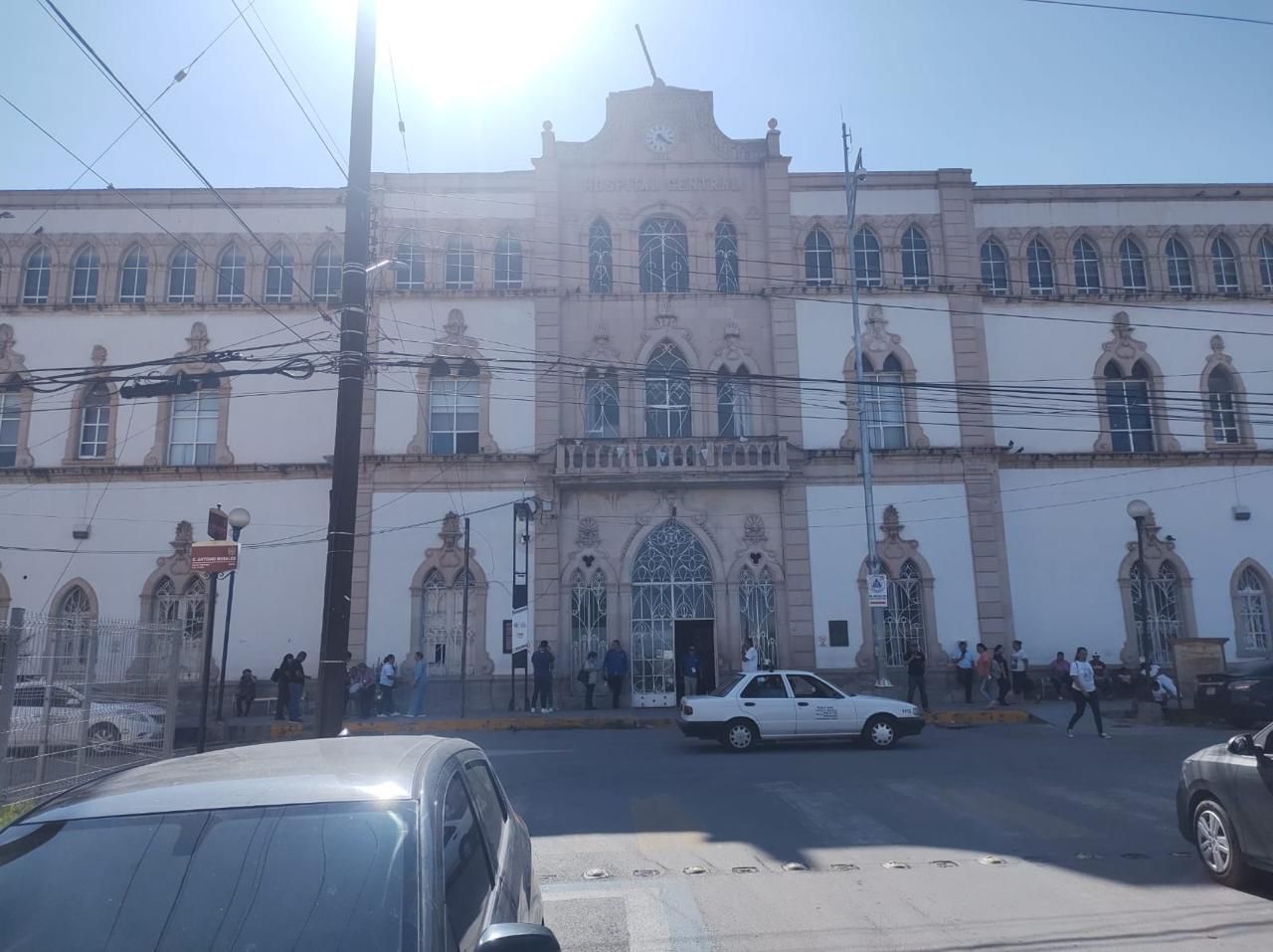 Vista exterior de un gran edificio ornamentado con entrada central. Coches y peatones al frente.