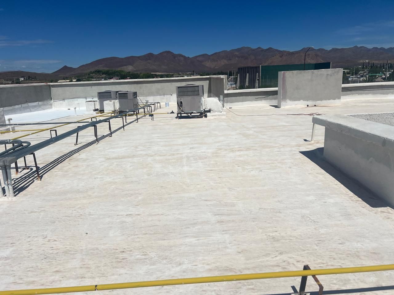 Techo blanco con unidades HVAC, barandillas amarillas y montañas distantes bajo un cielo azul.