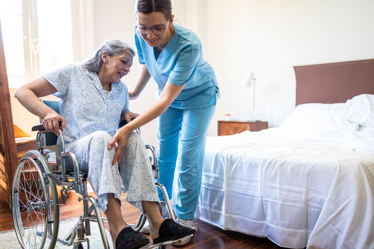 A geriatric nursing assistant helping a senior woman get out of her wheelchair and move to her bed.