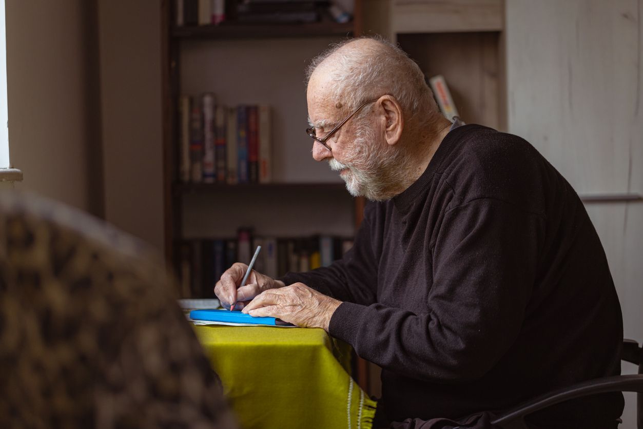 A senior, possibly living with dementia, writing something or signing documents in his living area