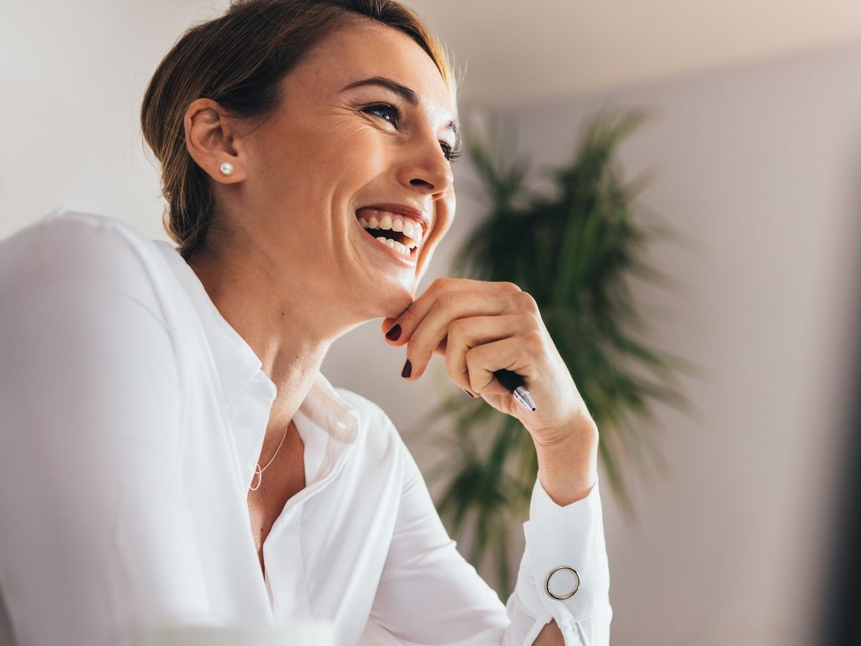 A woman is smiling while sitting at a desk with a pen in her hand.