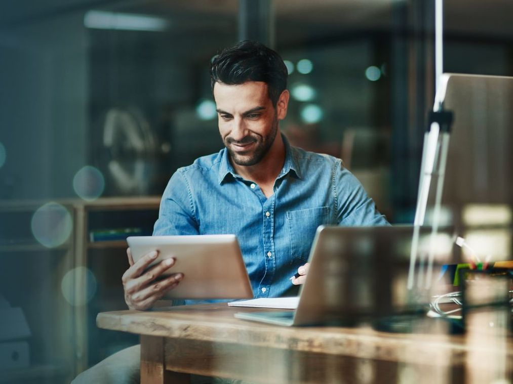 A man is sitting at a desk using a tablet and a laptop.