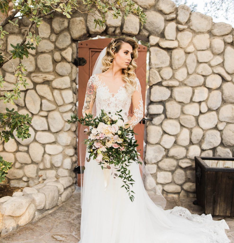 A bride in a wedding dress is standing in front of a stone wall holding a bouquet of flowers.
