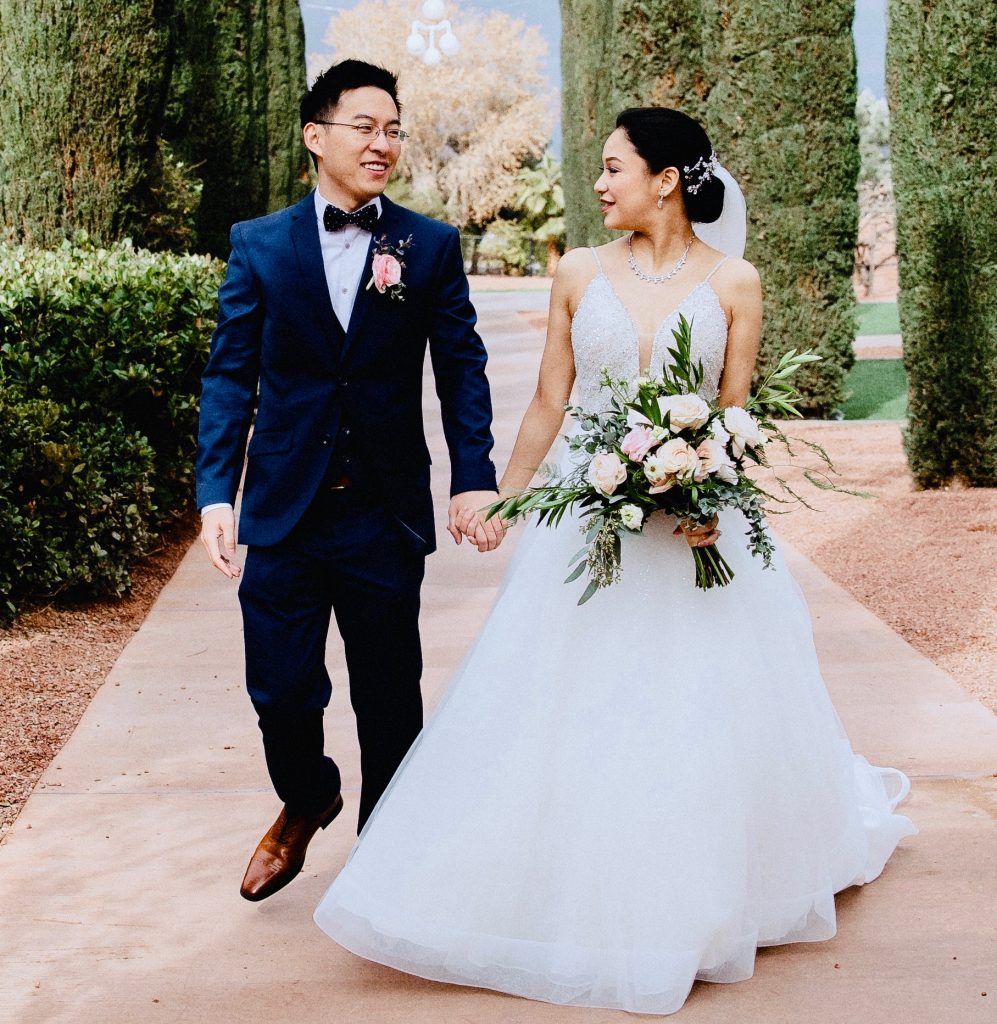 A bride and groom are walking down a path holding hands
