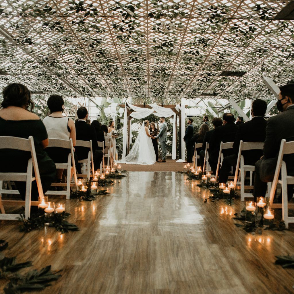 A bride and groom are getting married in a greenhouse surrounded by candles and leaves.