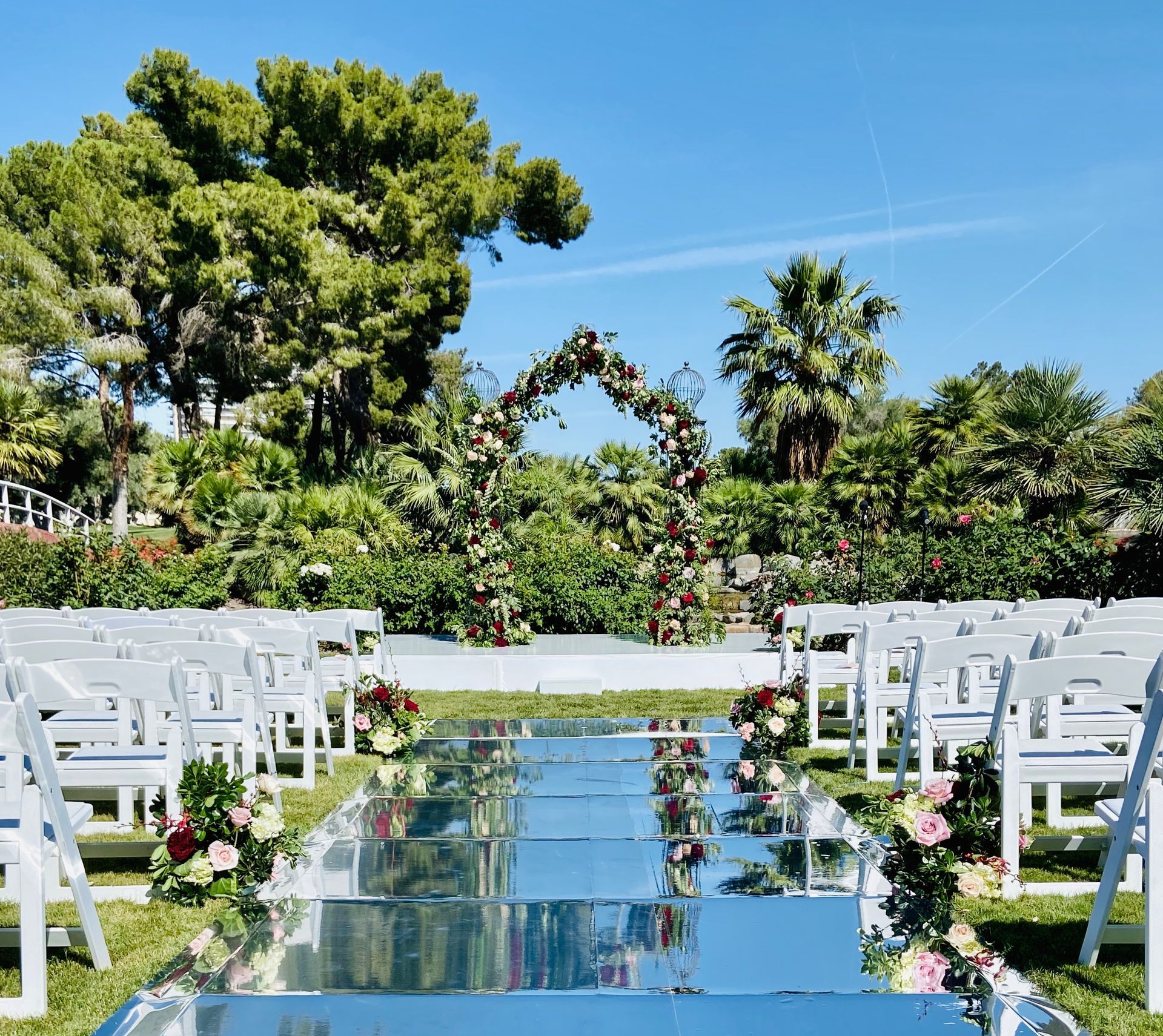 A row of white chairs are lined up in front of a mirrored aisle decorated with flowers.