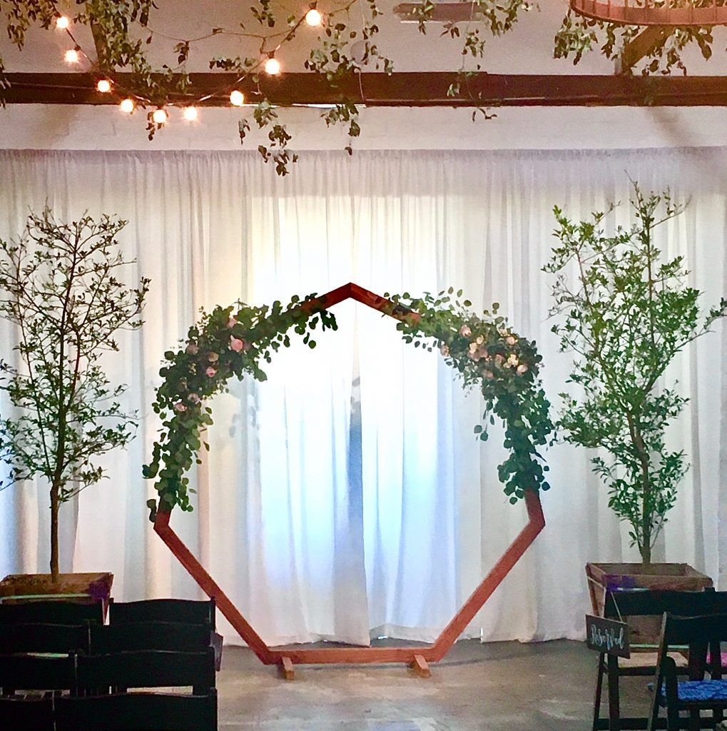 A wooden arch decorated with flowers and leaves is in front of a white curtain.