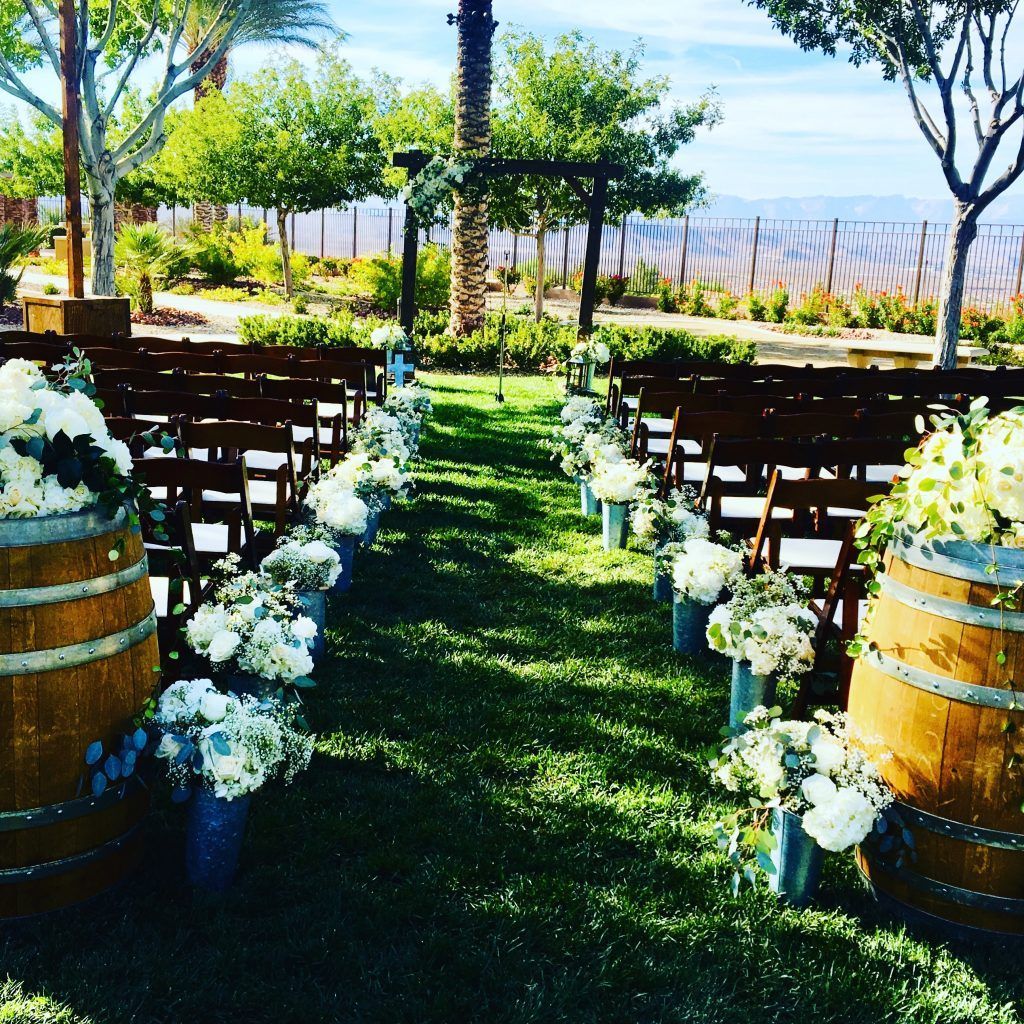 A row of wooden chairs lined up on a lush green field surrounded by barrels and flowers.