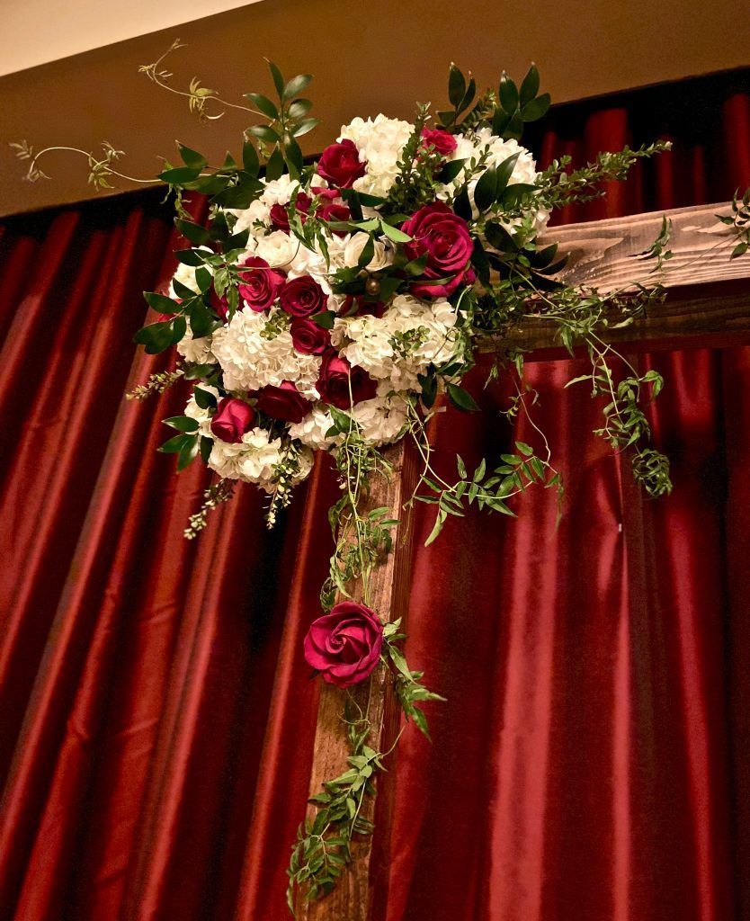 A wooden arch decorated with red and white flowers is sitting in front of a red curtain.