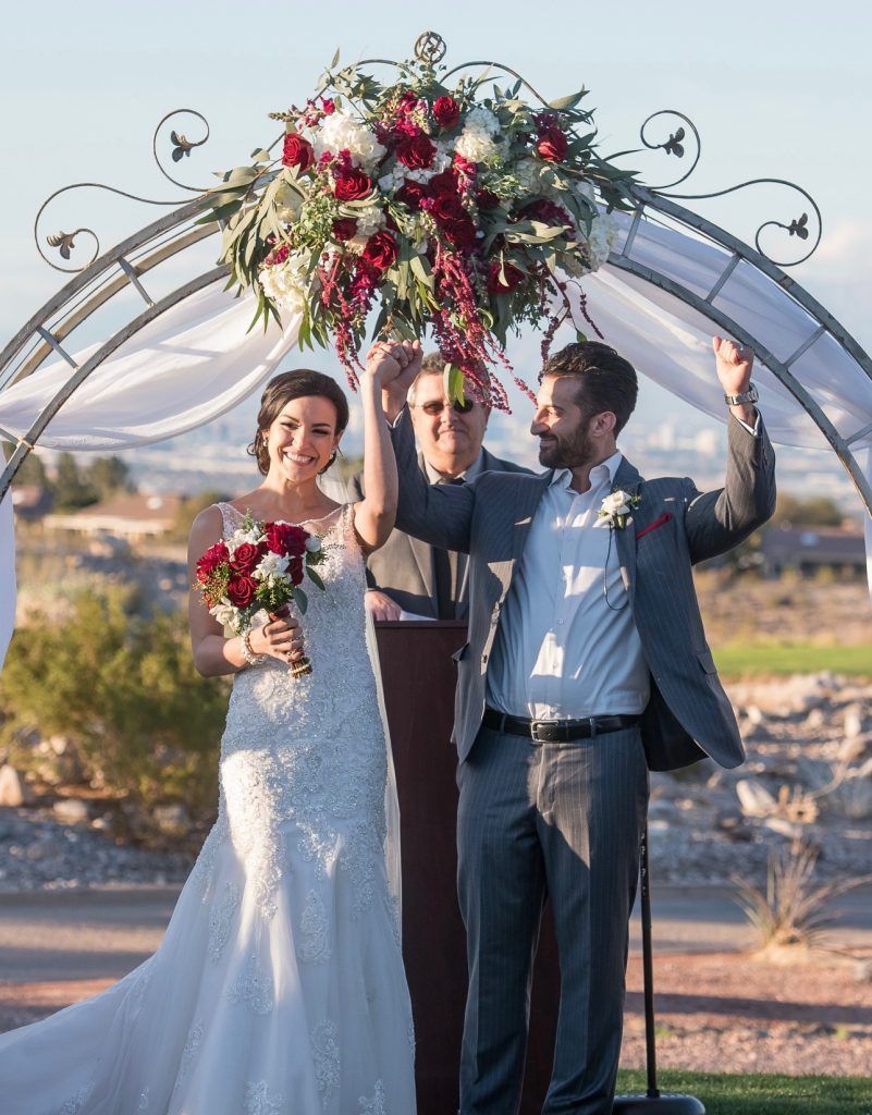 A bride and groom are standing under a floral arch at their wedding ceremony.