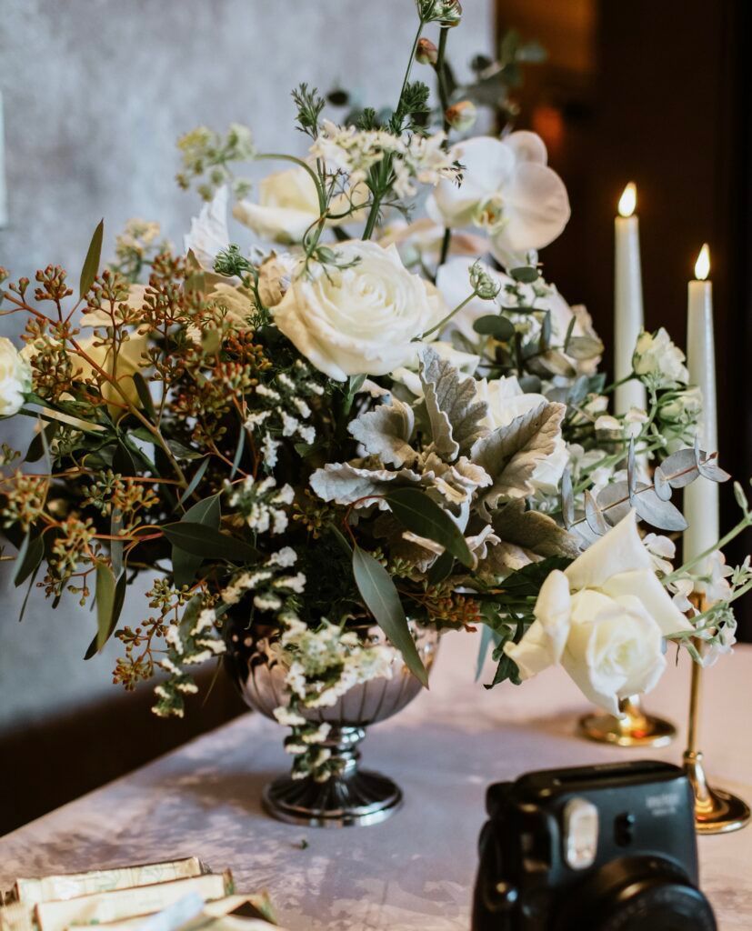 A vase filled with white flowers and candles on a table next to a camera.
