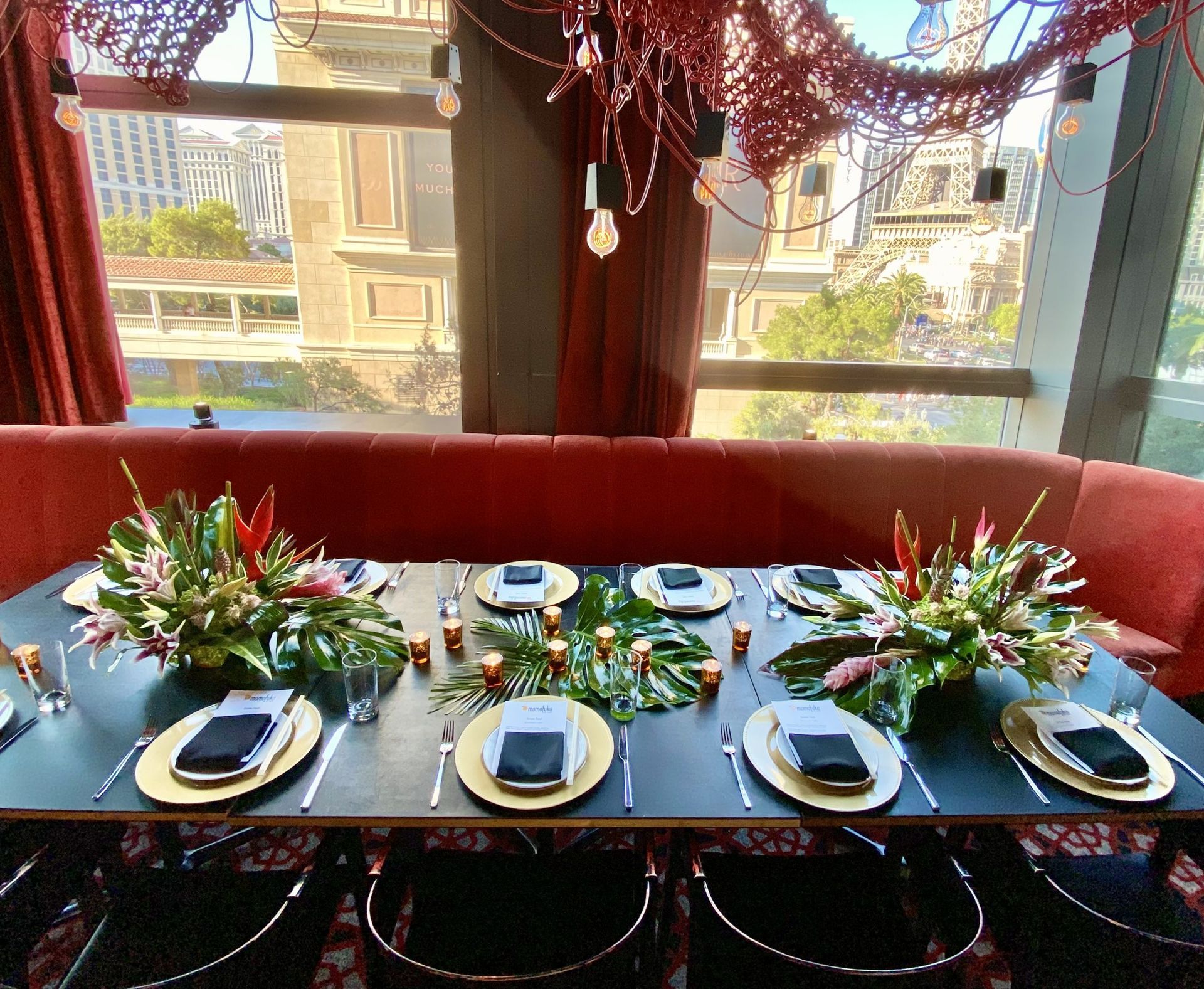 A long table with plates , napkins , utensils and flowers on it in front of a window.