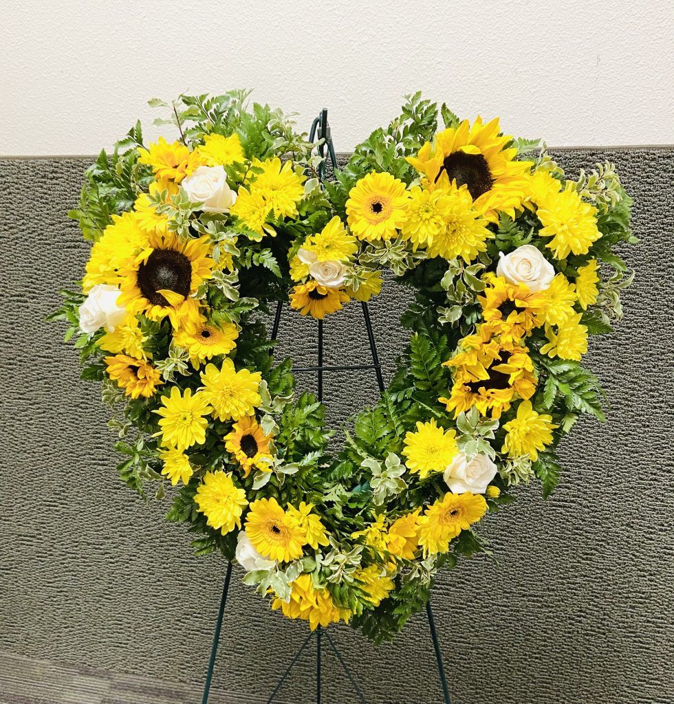 A heart shaped wreath made of yellow flowers is sitting on a stand.