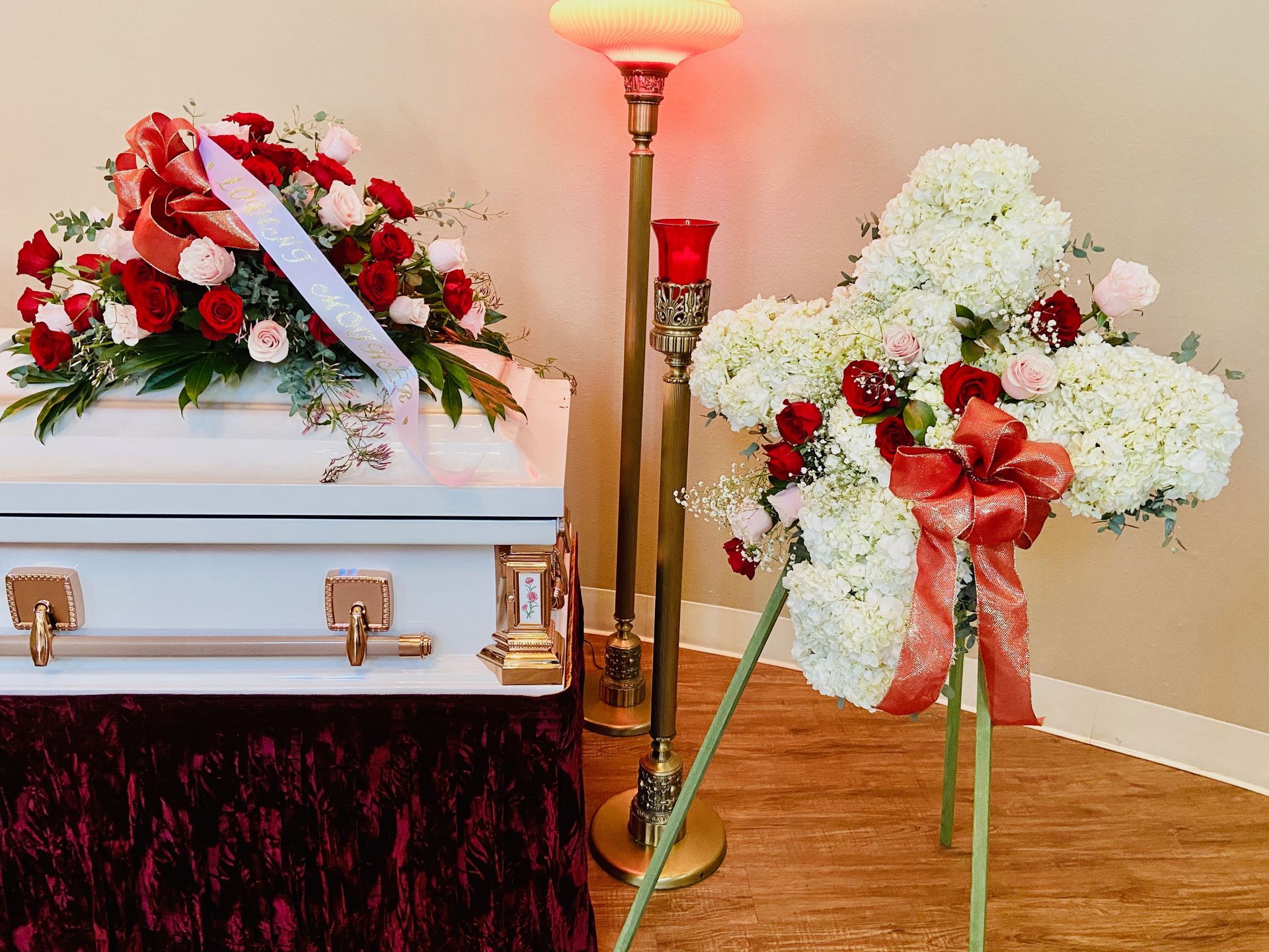 A white coffin is sitting next to a wreath of red and white flowers.